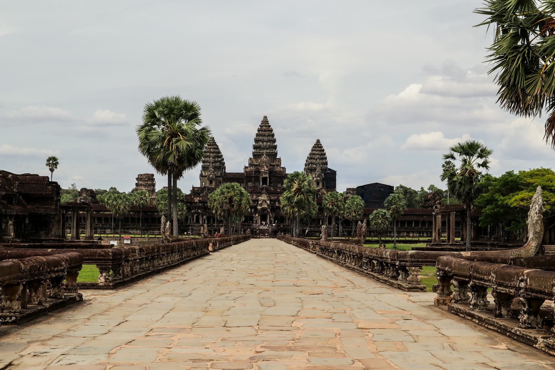 The long stone causeway leads toward the towers of Angkor Wat, lined with balustrades and palm trees under a cloudy sky.