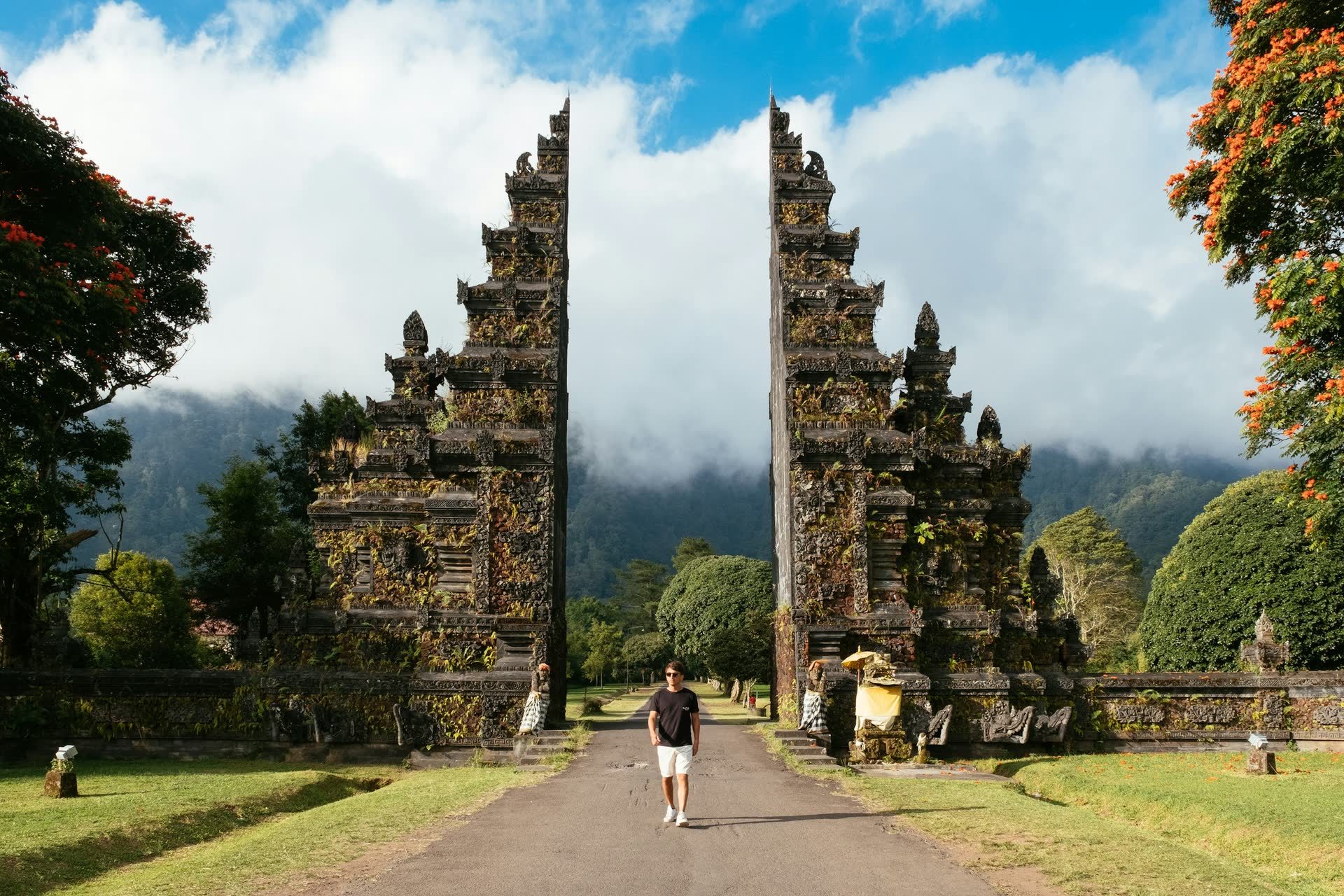 A road runs through the split stone Handara Gate in Bali, framed by trees and misty mountains under a blue sky.