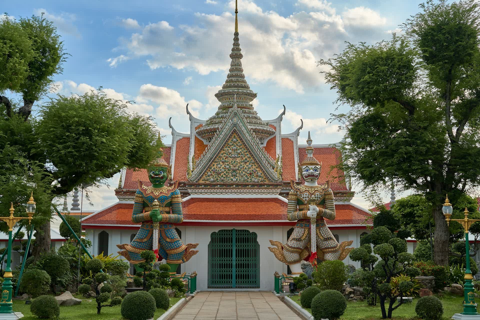 Two giant guardian statues flank the gateway to the ordination hall at Wat Arun, with a path leading to its green door under a bright sky.