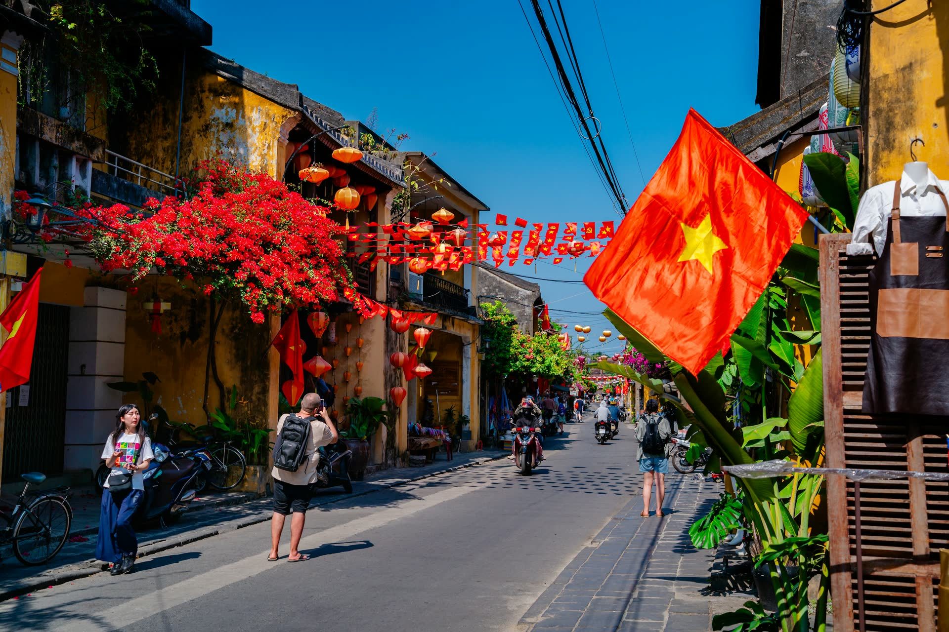 A sunlit street in Hội An's old town, strung with red lanterns and bougainvillea, with a Vietnamese flag in the foreground.